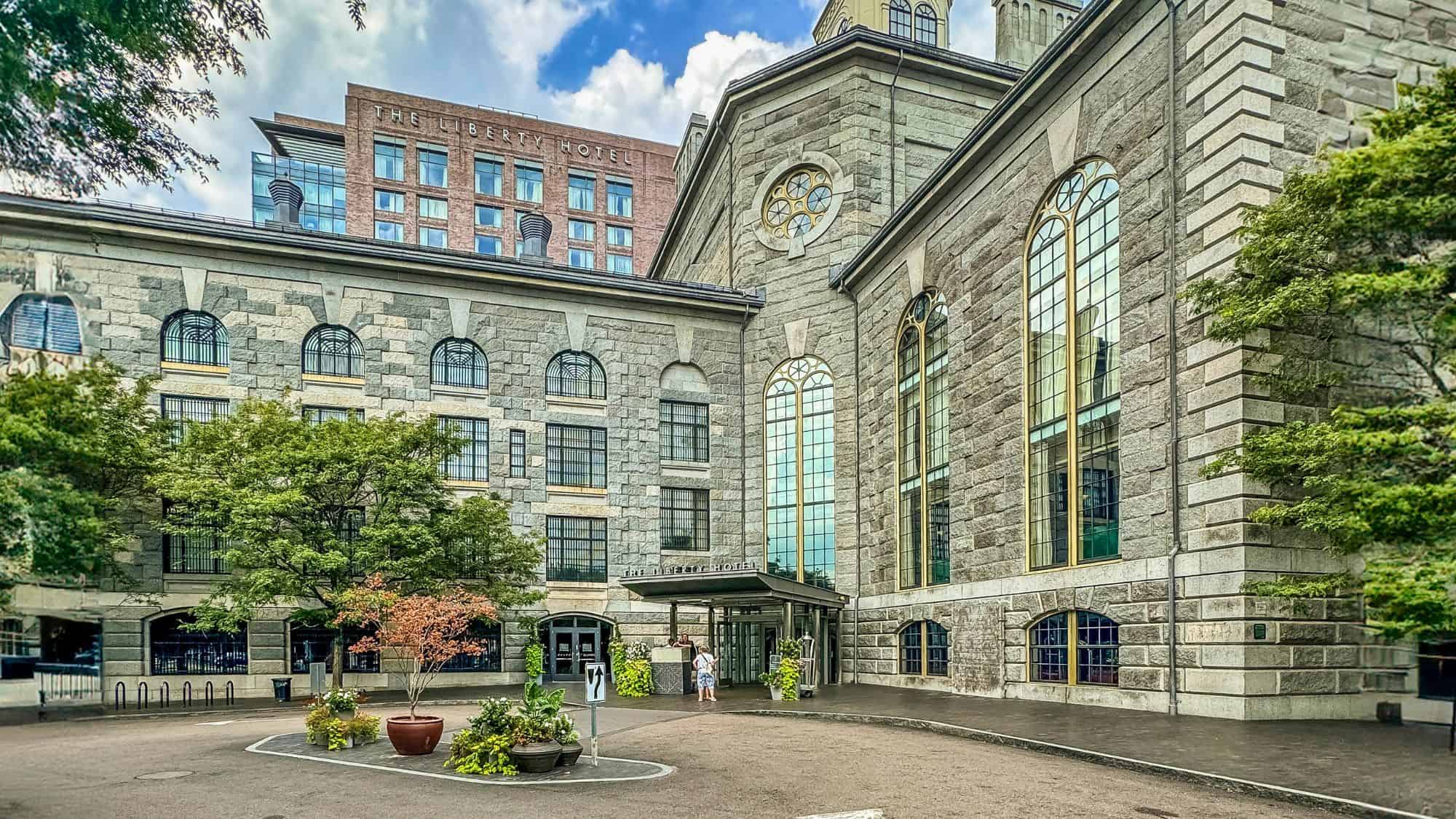 The Liberty Hotel in Boston features a historic stone façade with tall arched windows, surrounded by leafy trees, with the modern hotel sign and red brick tower visible behind it.
