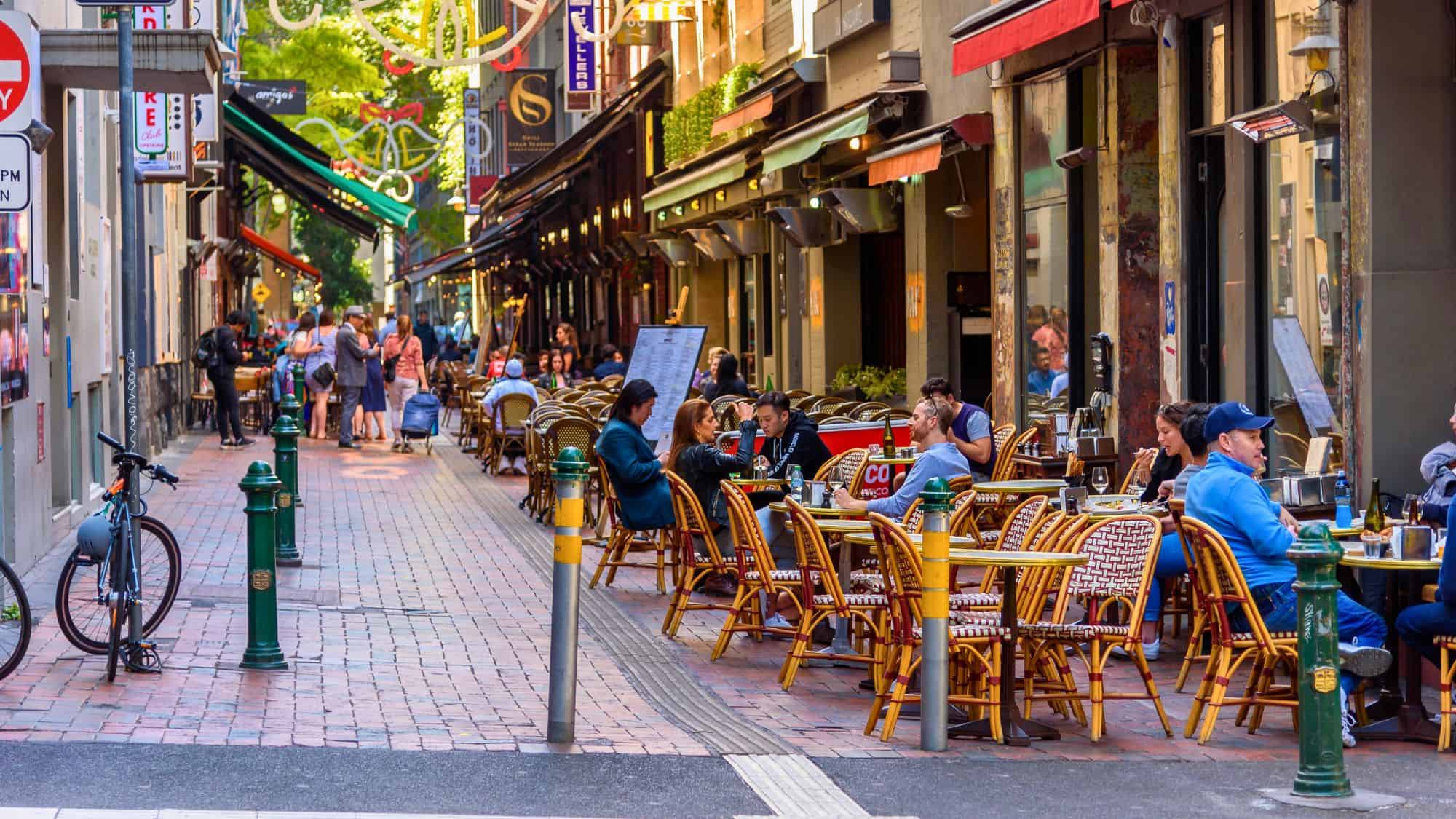 Narrow laneway in Melbourne lined with bustling outdoor cafés and bistros, offering a cozy and colorful urban dining atmosphere.