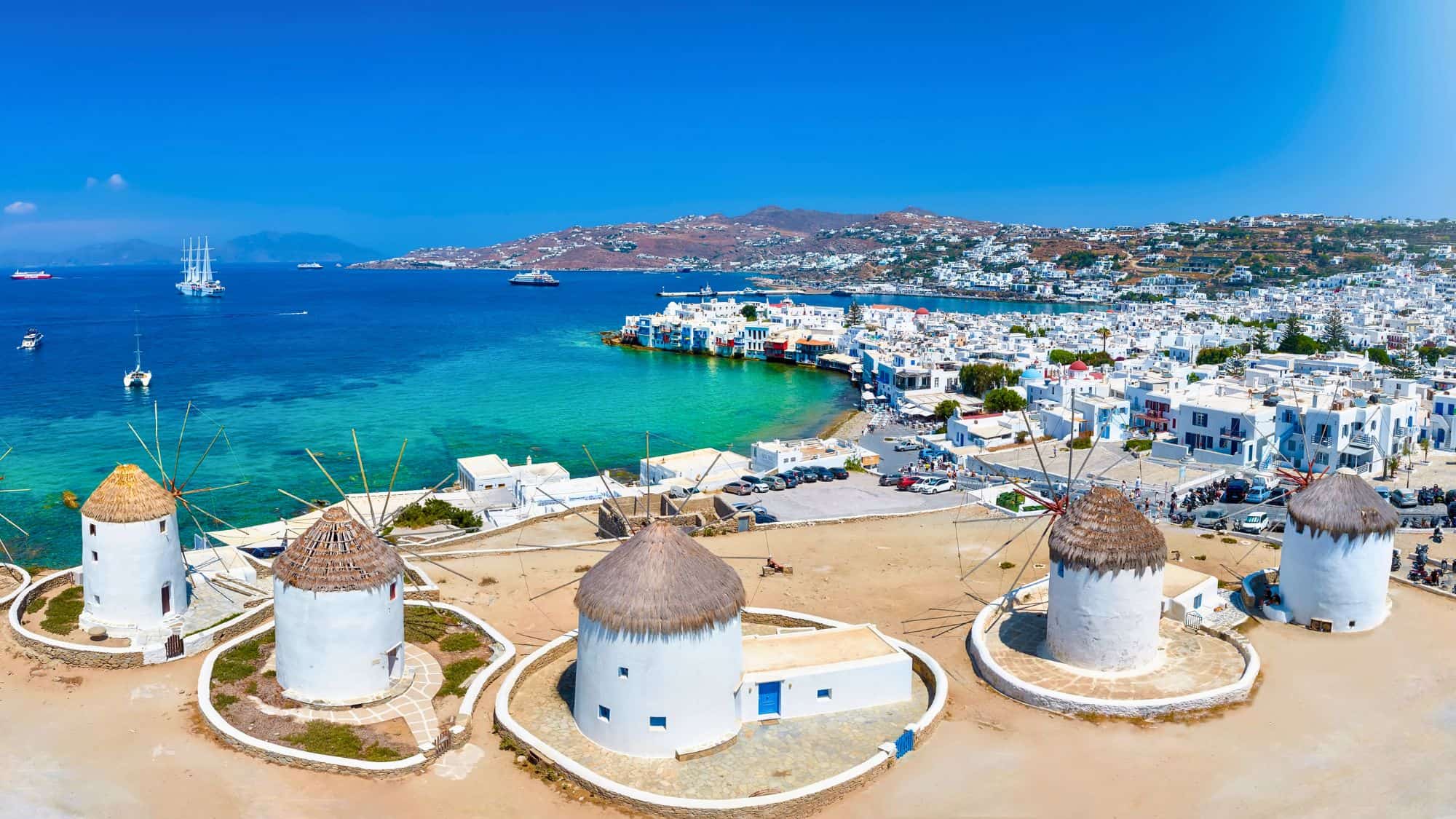 Traditional whitewashed windmills with thatched roofs overlook the Aegean Sea and the charming coastal town of Mykonos, Greece, under a bright blue sky.