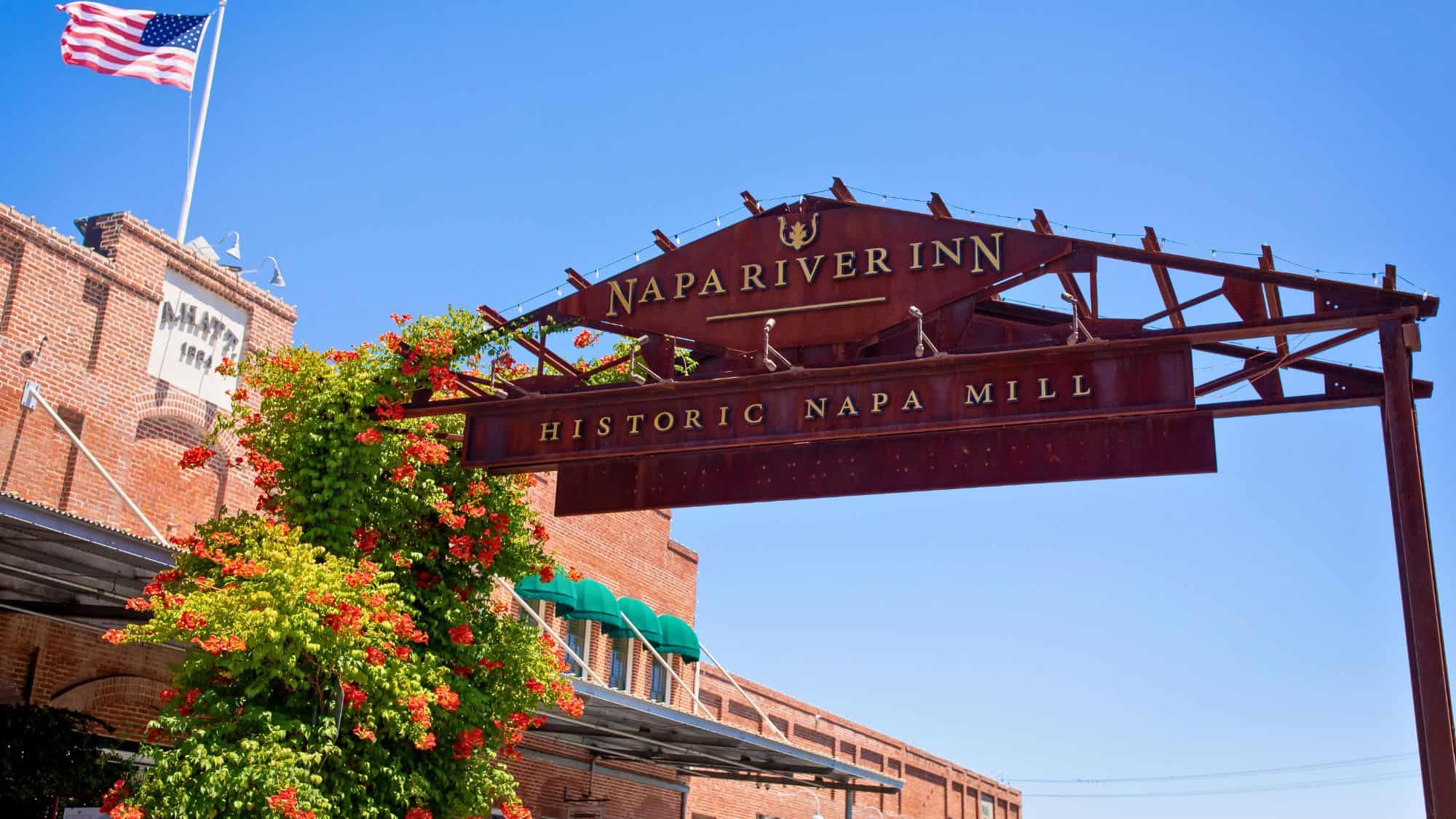 A rustic metal archway with "Napa River Inn" and "Historic Napa Mill" signage stretches over a walkway lined with blooming red and orange flowers and red-brick buildings.