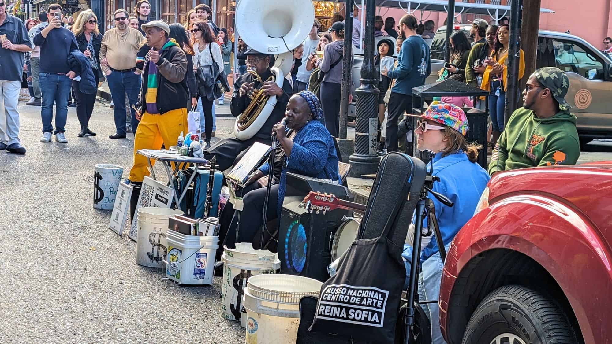 A lively group of musicians performs on a street corner in New Orleans, surrounded by buckets, instruments, and a crowd of captivated onlookers.