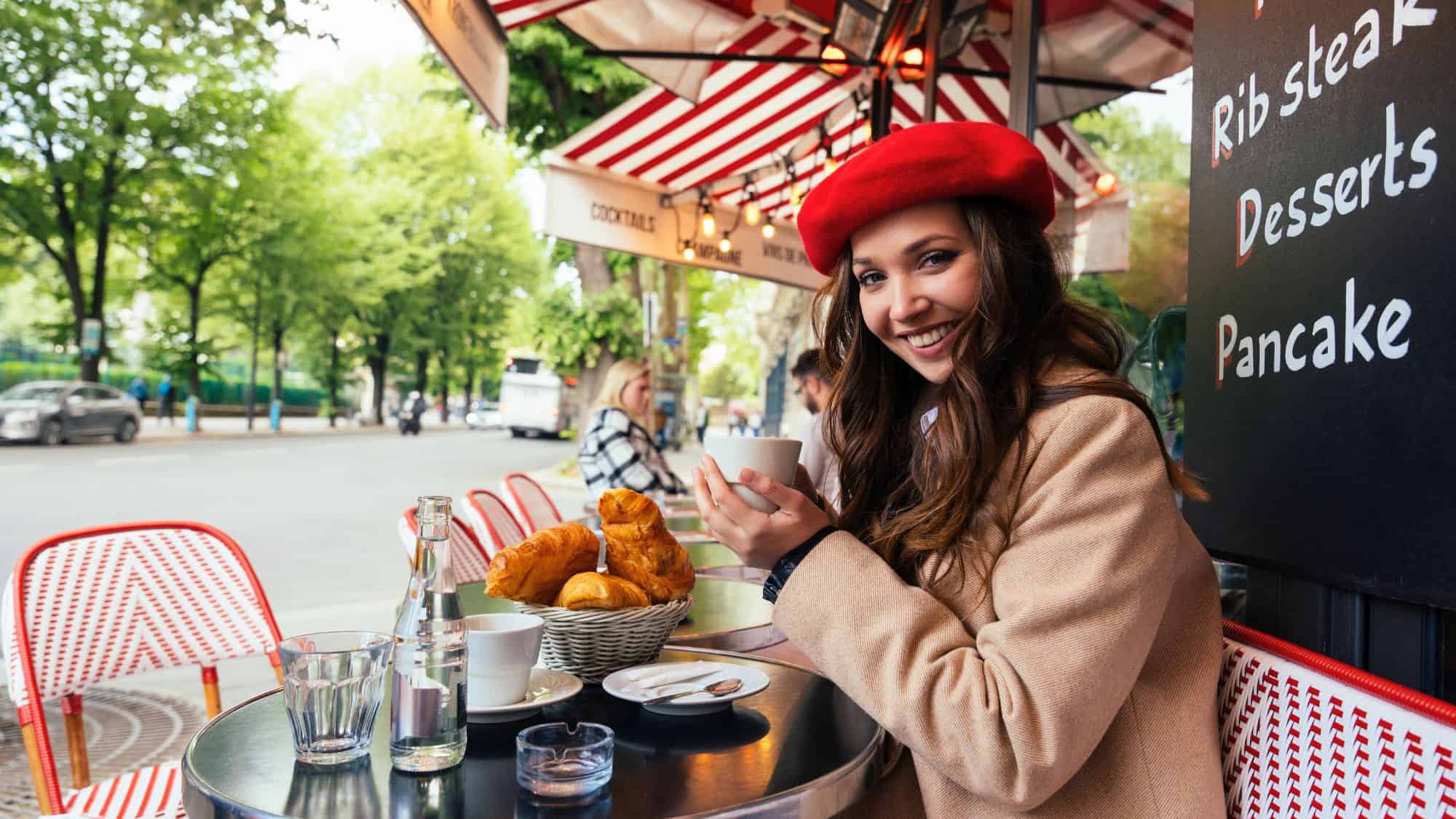 A cheerful woman in a red beret enjoys coffee and croissants at a classic Paris sidewalk café, with red-striped awnings and cozy seating.