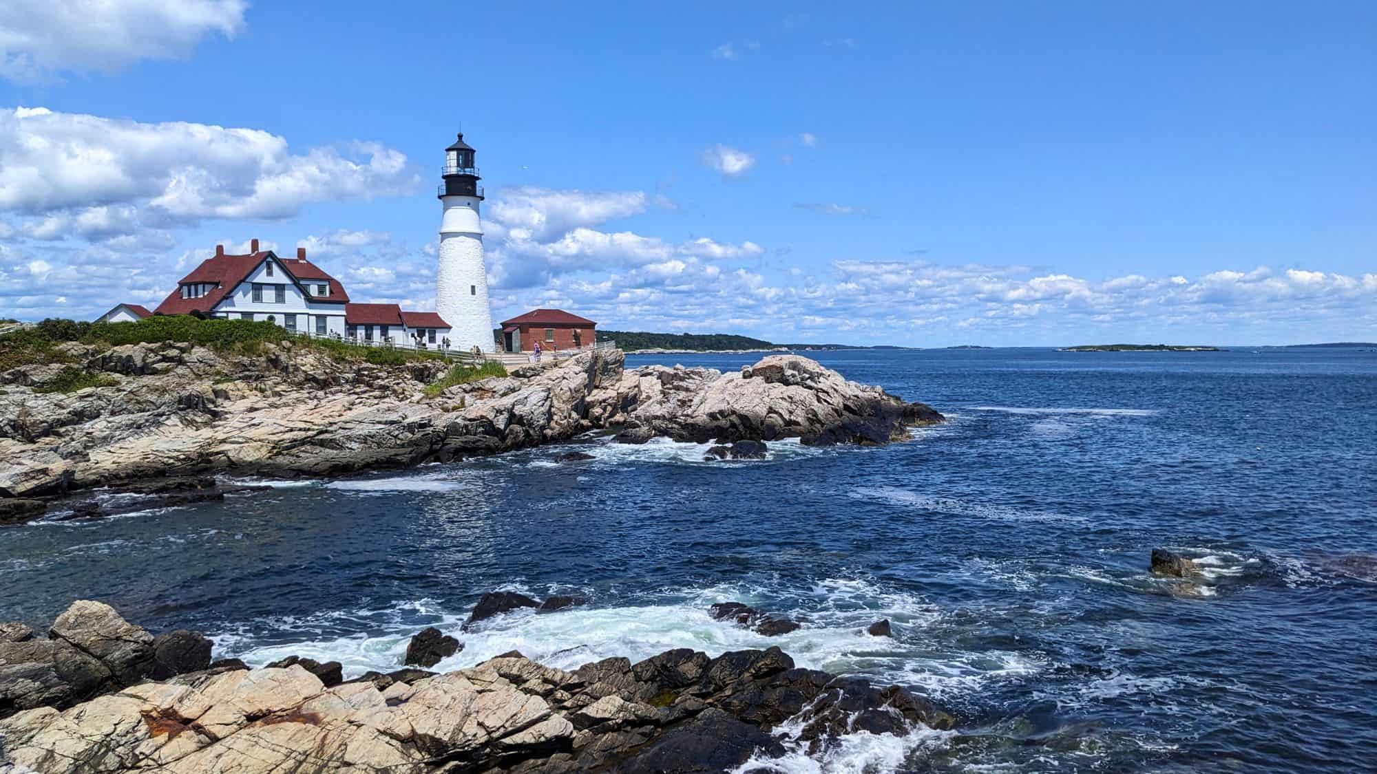 The iconic Portland Head Light stands on a rocky shoreline under a blue sky, with ocean waves crashing and historic white buildings nearby.