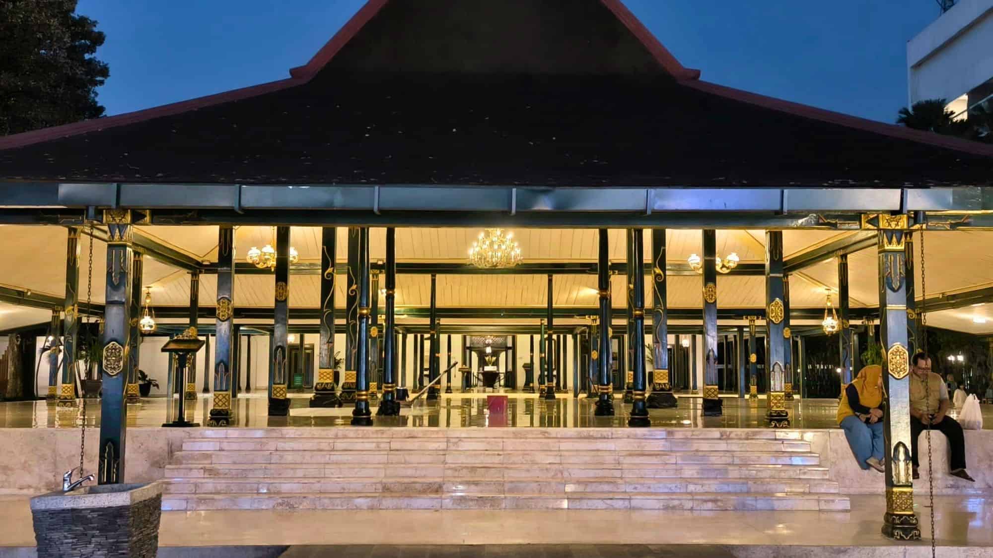An open-air Javanese-style pavilion with green and gold ornate pillars and chandeliers sits at the center of the Kraton Yogyakarta complex, with two people seated on the marble steps at night.