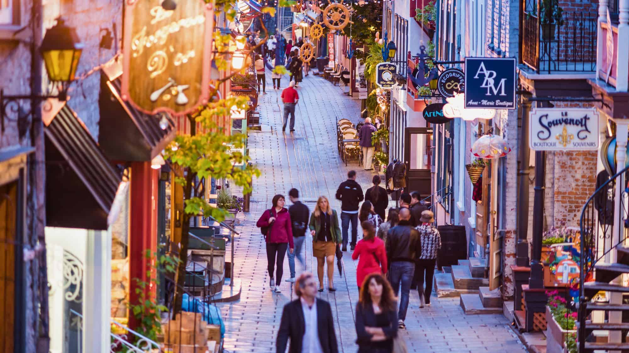 A cobblestone pedestrian street in Old Quebec is lined with boutique shops and warm lights as people stroll past hanging signs and cozy storefronts in the early evening.