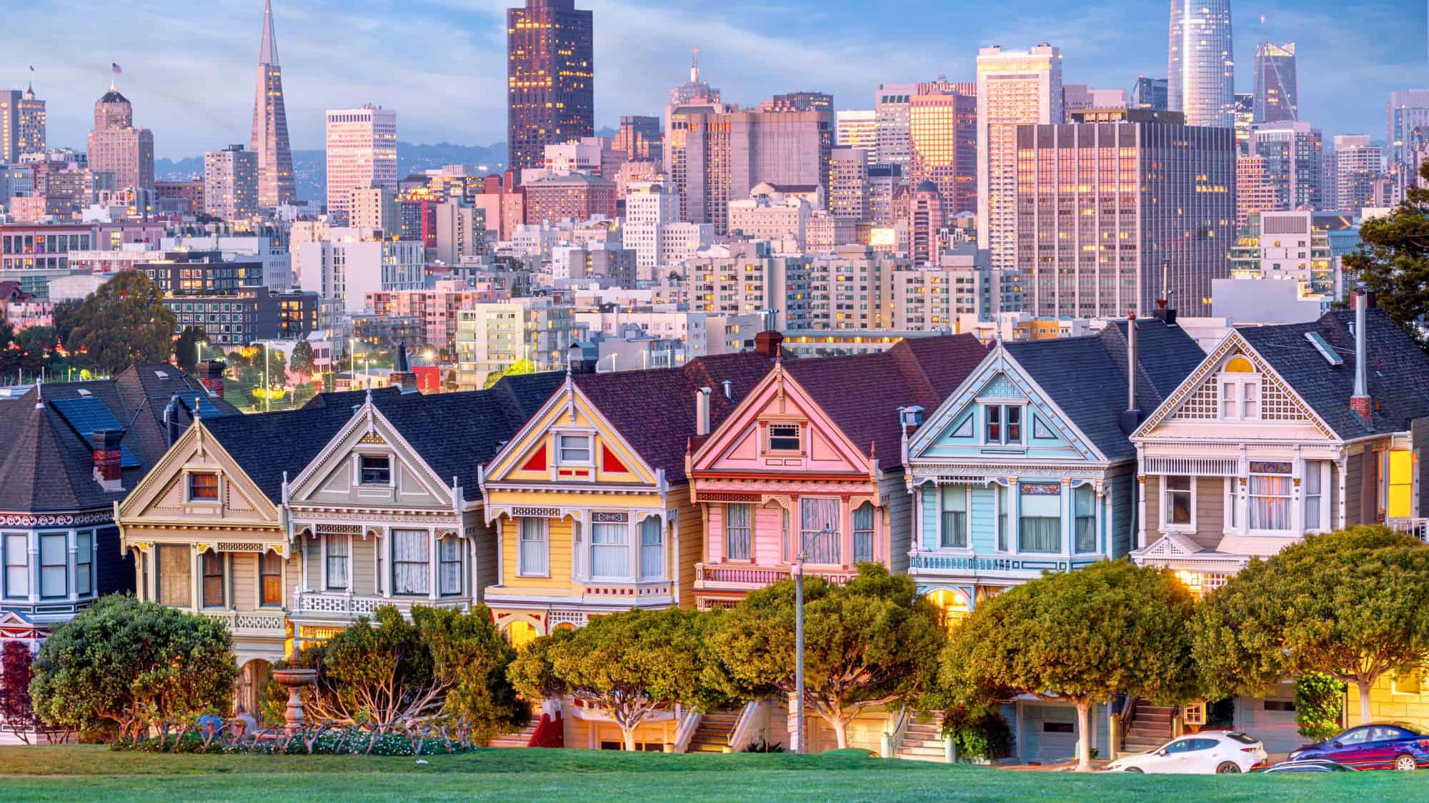 The iconic row of colorful Victorian houses known as the Painted Ladies sits in front of the modern San Francisco skyline at sunset.