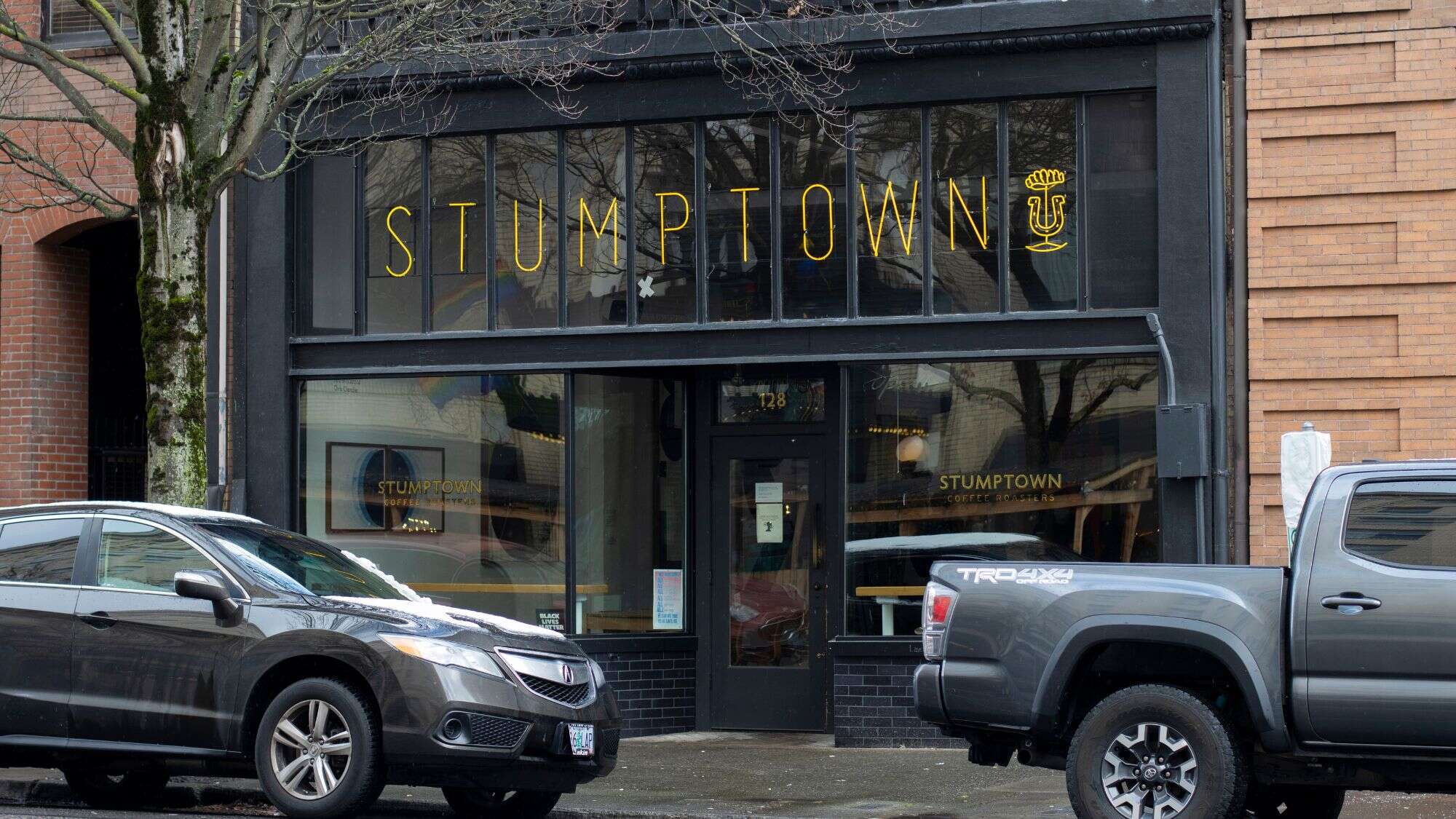 The iconic black façade of Stumptown Coffee Roasters in Portland, Oregon, sits between brick buildings, framed by parked cars and a rainy street.