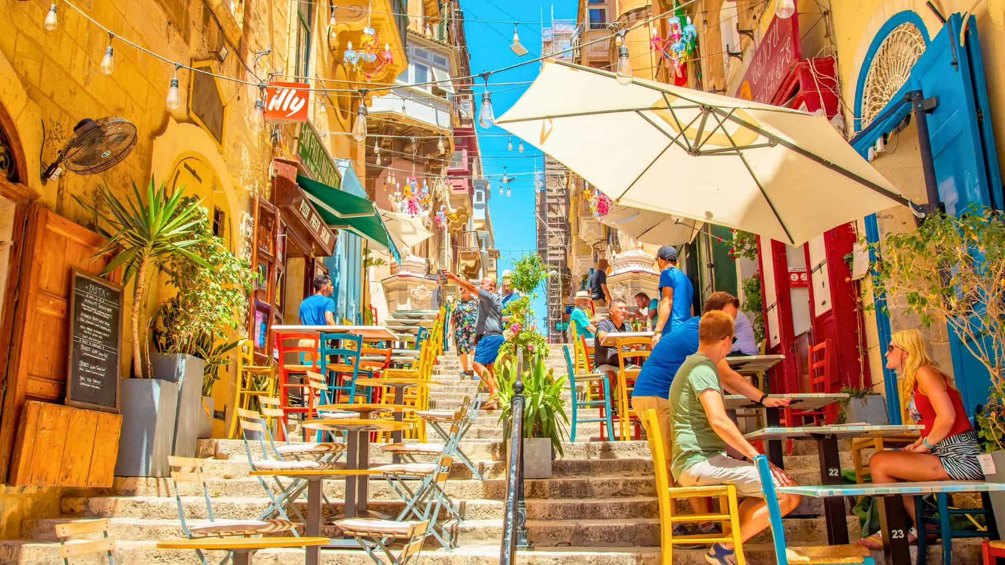 Outdoor cafés with bright chairs and umbrellas line a sunlit stone staircase in Valletta, Malta, creating a vibrant and inviting atmosphere for dining and conversation.