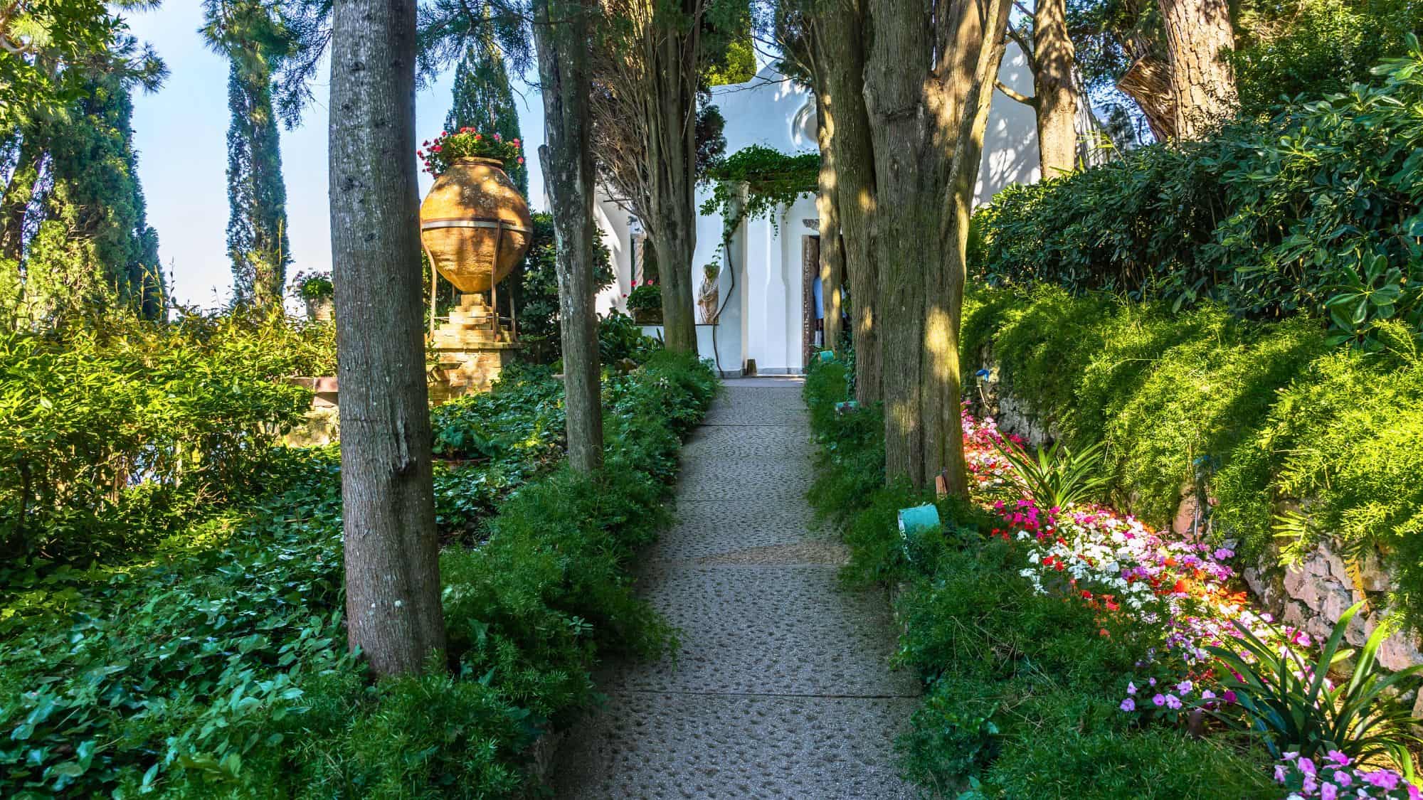 A stone pathway flanked by tall trees and lush greenery leads to a white building, with colorful flowers and a large terracotta urn adding charm to the serene garden setting.