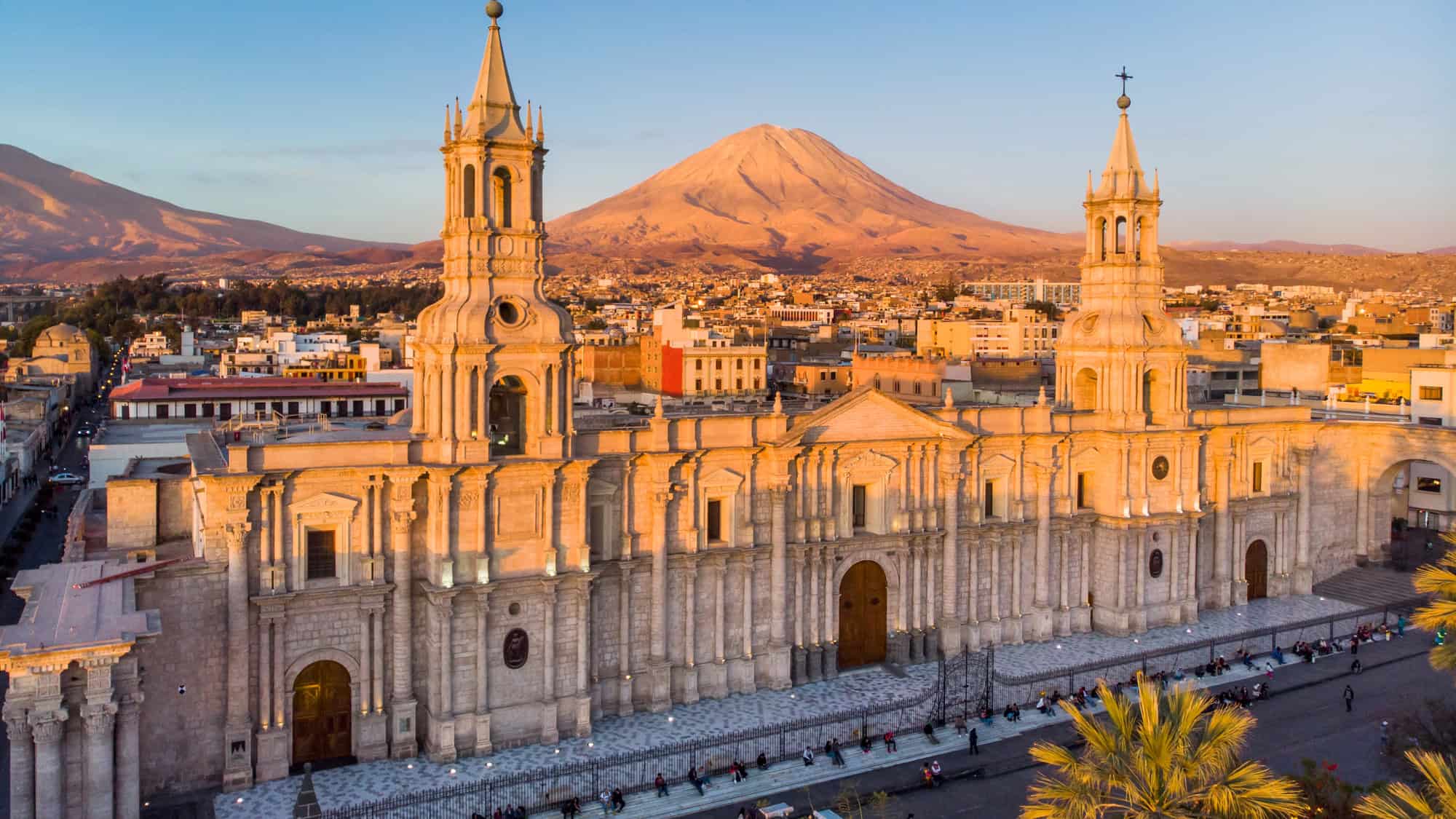 Arequipa’s white stone cathedral dominates the foreground with elegant twin towers, while the perfectly conical Misti Volcano looms in the distance under golden hour sunlight.
