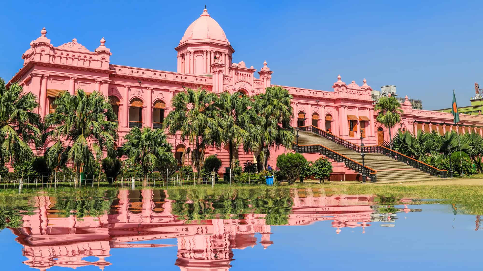 The vibrant pink Ahsan Manzil, once a royal palace in Dhaka, reflects in a still water feature, framed by palm trees and a clear blue sky.