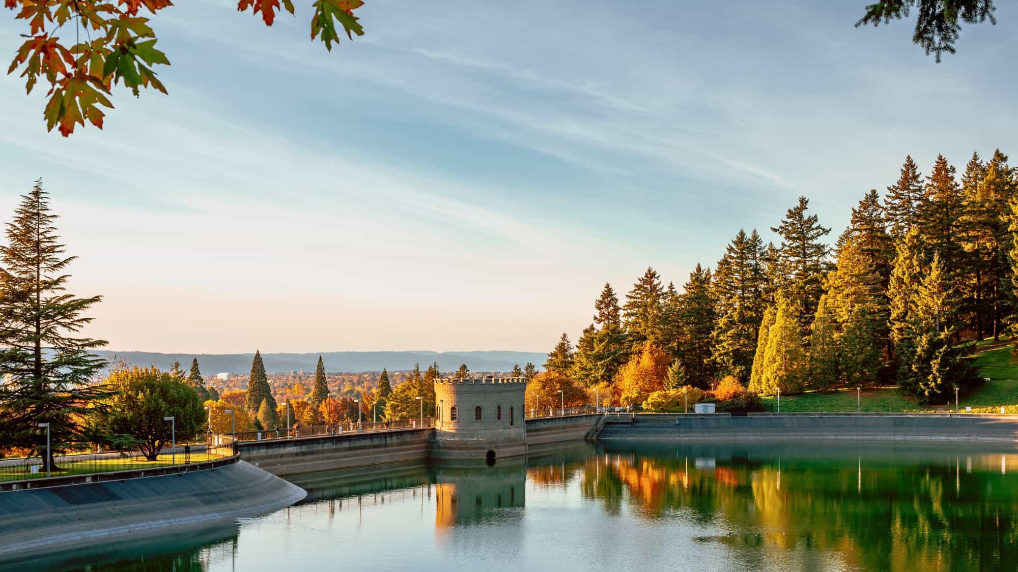 A stone gatehouse reflects in the still water of a tree-lined reservoir in Portland’s Mount Tabor Park, glowing with golden fall leaves under soft evening light.