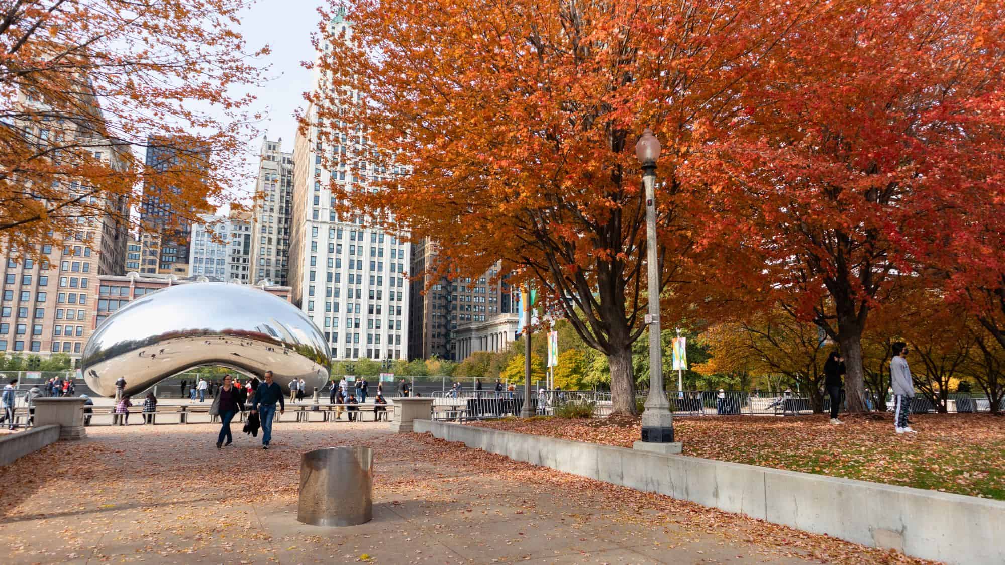 Visitors stroll through Chicago’s Millennium Park in fall, with the reflective Bean sculpture and vibrant red-orange trees standing out against surrounding skyscrapers.