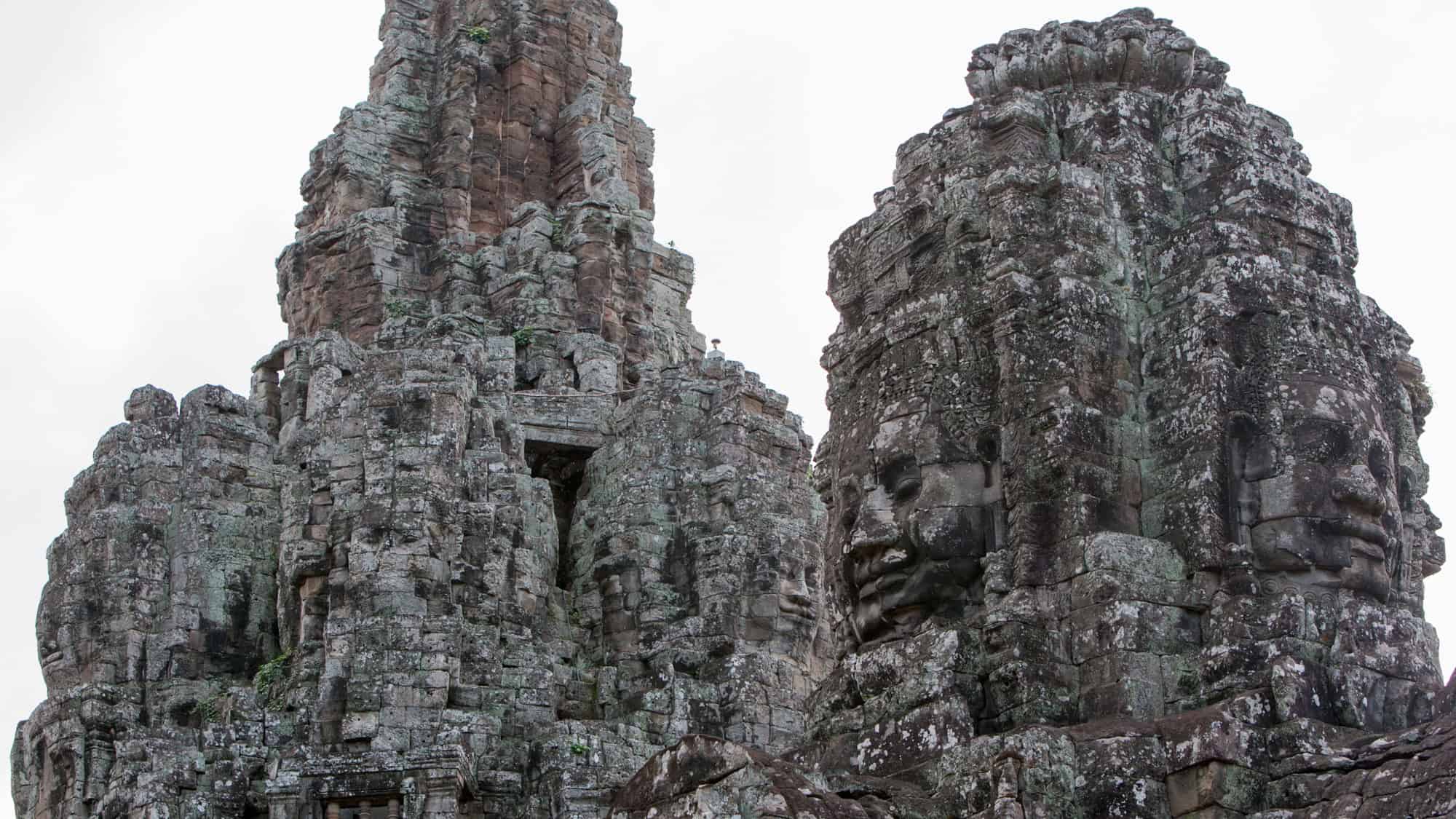 Massive stone towers at Cambodia’s Bayon Temple feature intricately carved serene faces, weathered by time and framed by a cloudy sky.