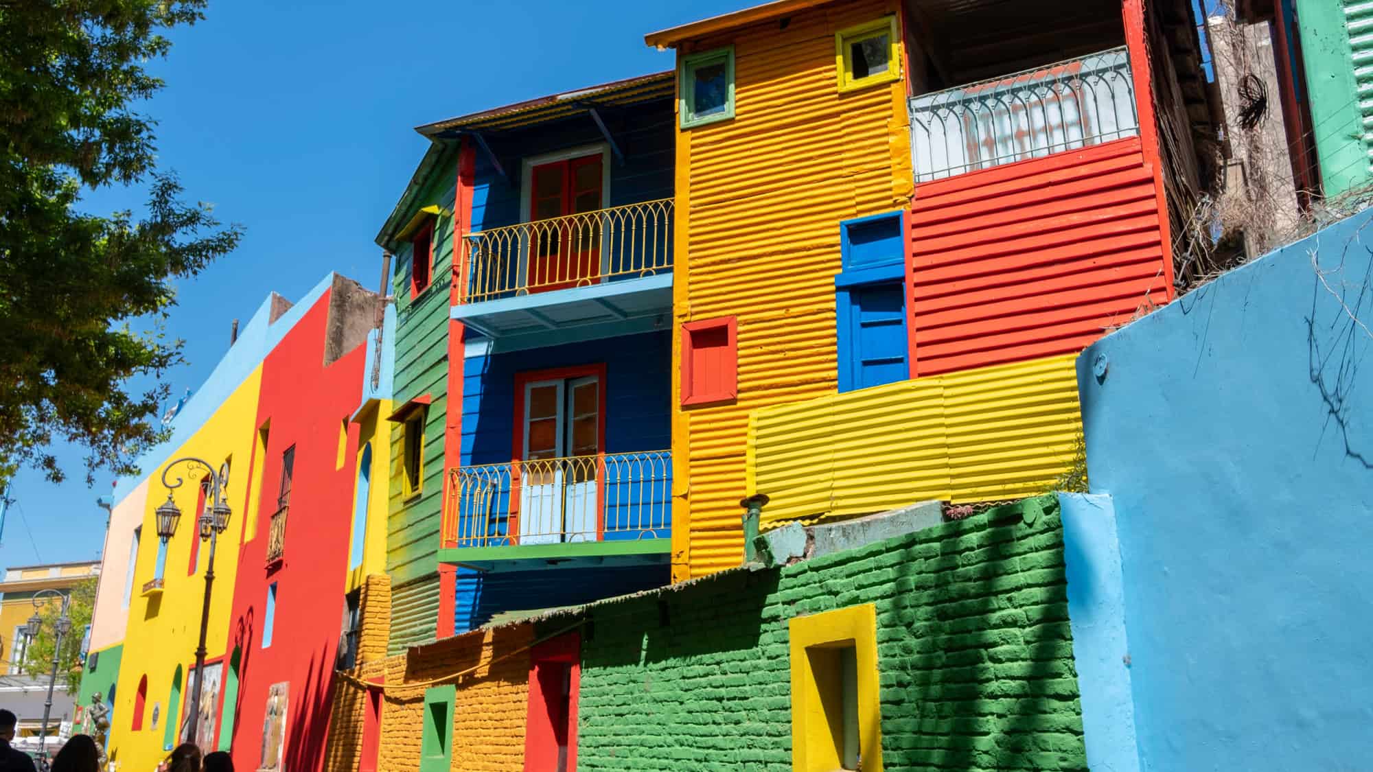 A row of vibrantly painted buildings in Buenos Aires’ La Boca neighborhood showcases bold reds, yellows, blues, and greens under a clear sky, with iron balconies and street art adding local flair.