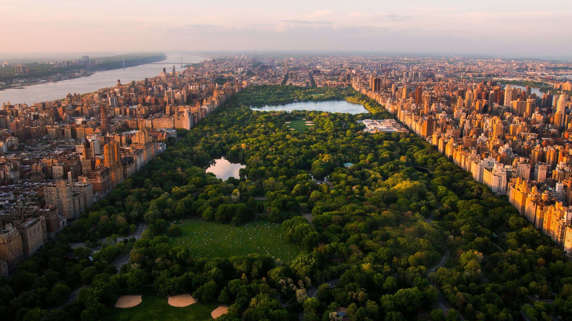 An aerial view of Central Park shows a green oasis stretching through the heart of Manhattan, flanked by dense rows of high-rise buildings glowing in the evening light.