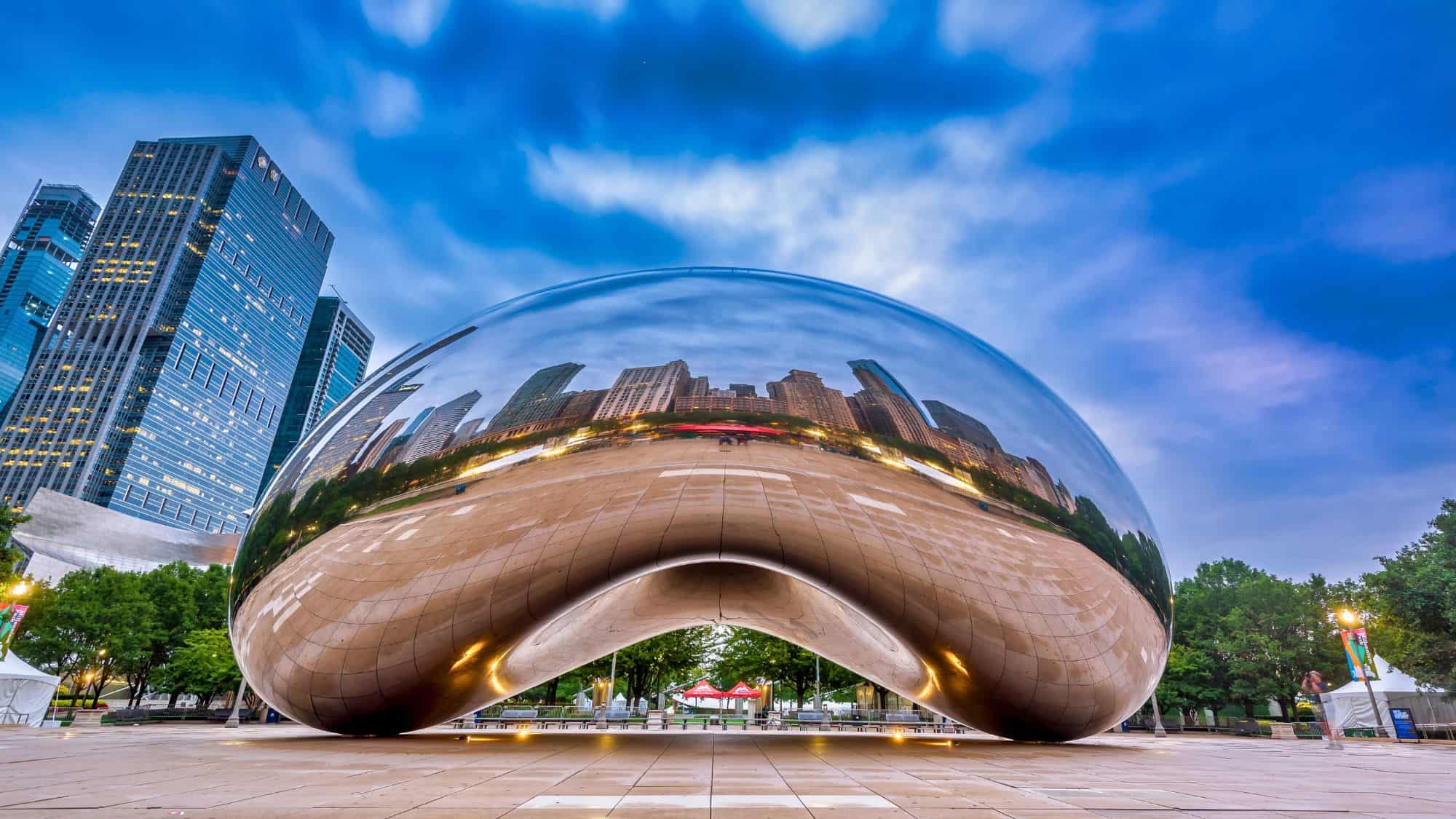 The reflective surface of Chicago’s Cloud Gate sculpture, also known as “The Bean,” mirrors the city skyline under a dramatic blue evening sky in Millennium Park.