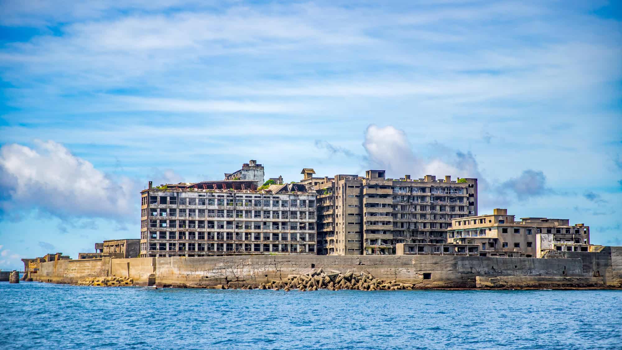 An eerie cluster of crumbling gray buildings rises from the sea on Hashima Island, also known as Battleship Island, under a bright blue sky.