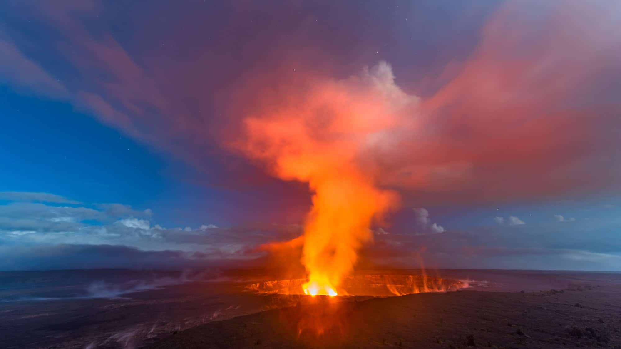 Molten lava glows intensely from a crater, sending up a towering plume of orange smoke under a twilight sky filled with pink and purple clouds.