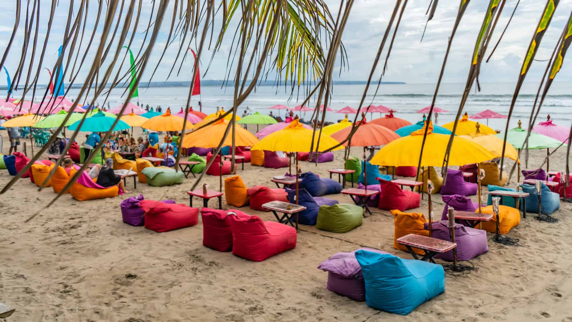 A lively beachfront in Bali is filled with bright beanbags and rainbow umbrellas, as people relax under tropical skies and swaying palms.
