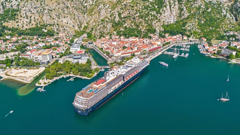 A large cruise ship is anchored in the scenic bay of Kotor, Montenegro, with the walled old town nestled at the base of steep green mountains.