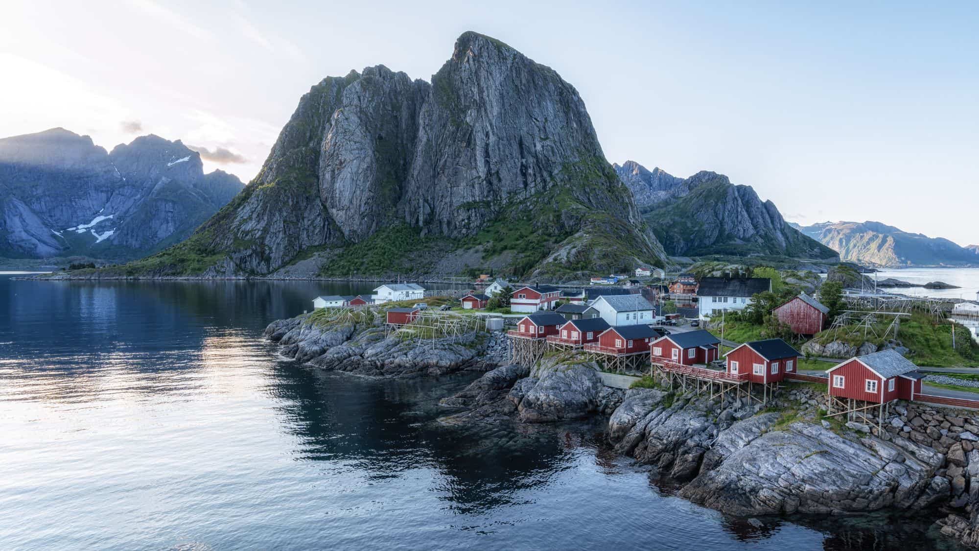 Charming red and white cottages sit on rocky outcrops beside a still fjord, dwarfed by a towering, rugged mountain in Norway’s Lofoten Islands.