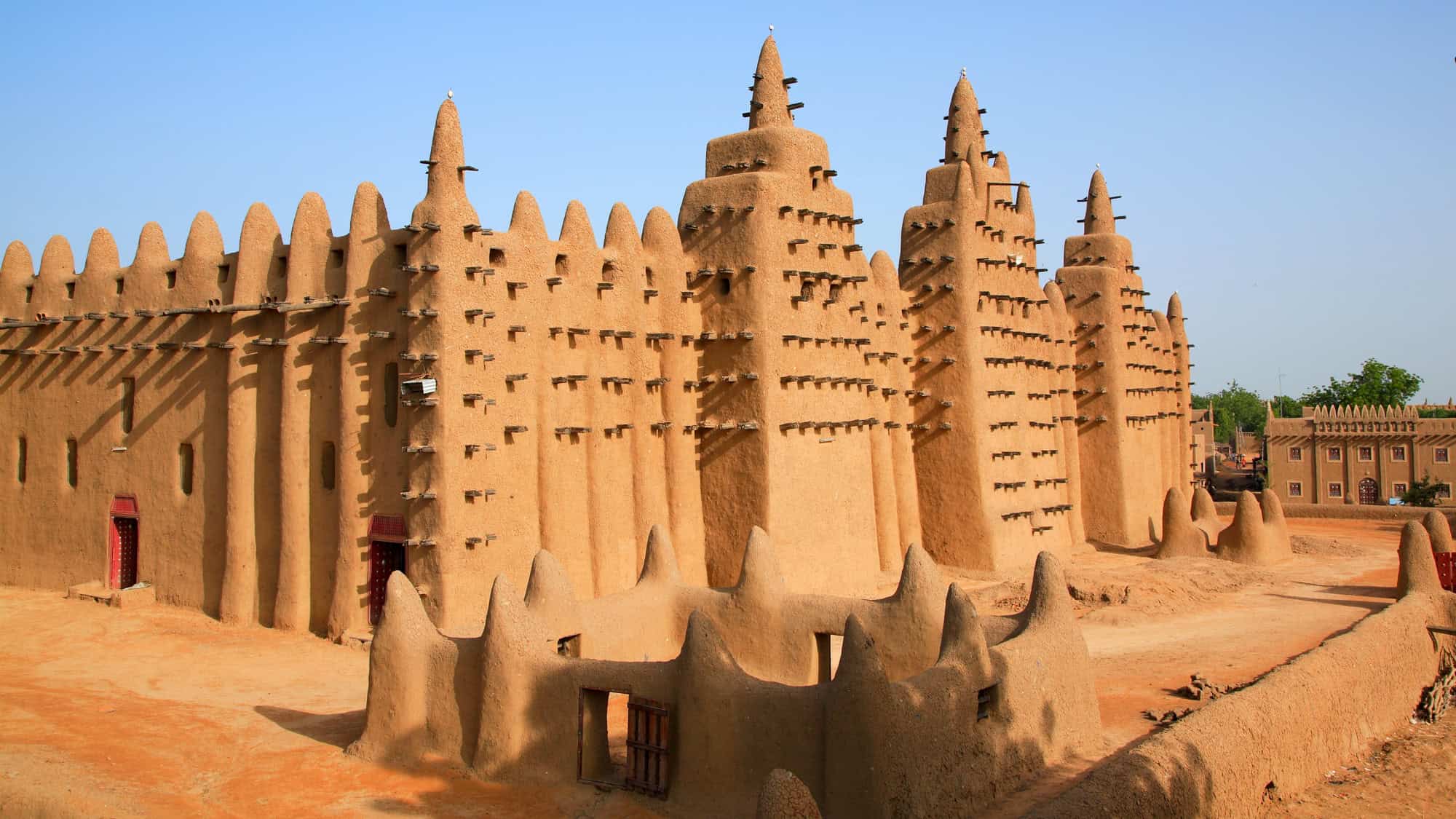 The towering adobe structure of the Great Mosque of Djenné, with its wooden scaffolding beams and conical spires, dominates the sandy landscape.