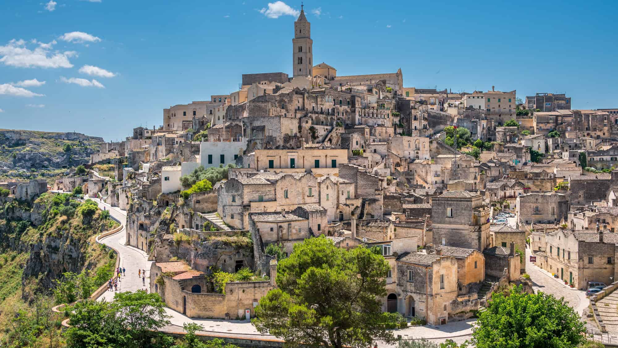 The ancient city of Matera in southern Italy is packed with stone buildings and cave dwellings cascading down a hillside, topped with a historic church tower.