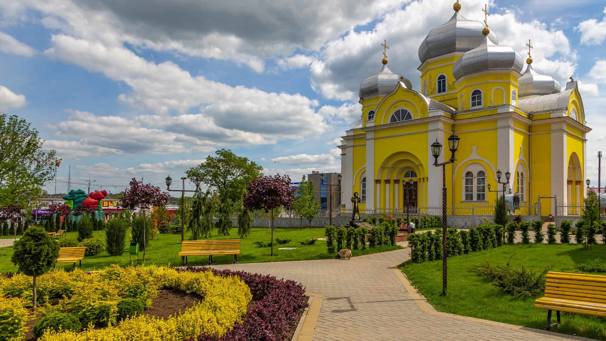 A bright yellow Orthodox church with silver domes stands beside a well-kept garden and cobblestone path, under a partly cloudy sky.