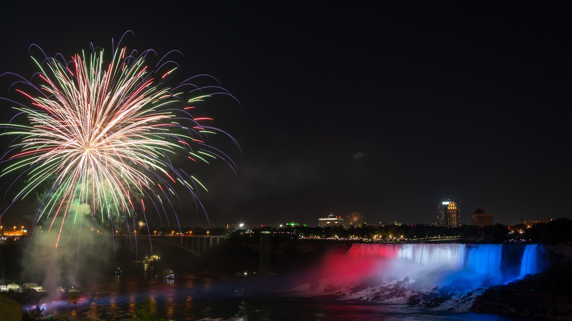 Bright, colorful fireworks burst over Niagara Falls at night while the falls are lit with red, white, and blue lights, creating a vibrant scene against the dark sky.