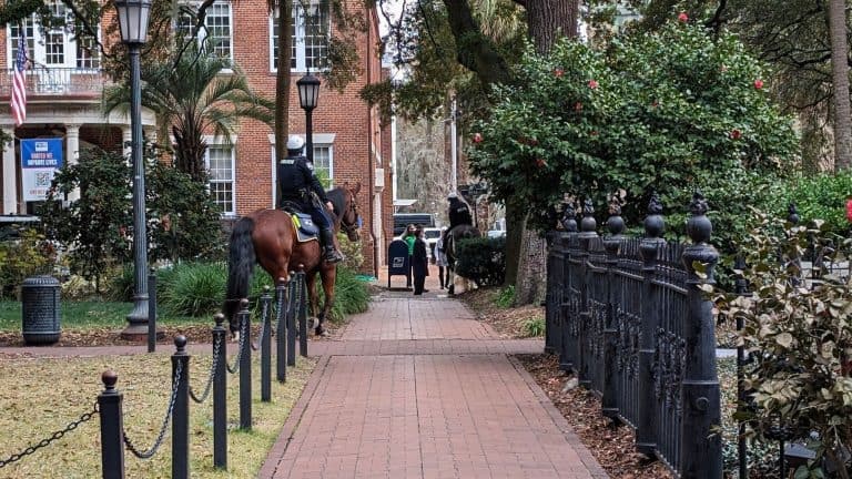 Two police officers on horseback patrol a quiet, brick-paved pathway lined with wrought iron fencing and lush greenery in historic downtown Savannah.