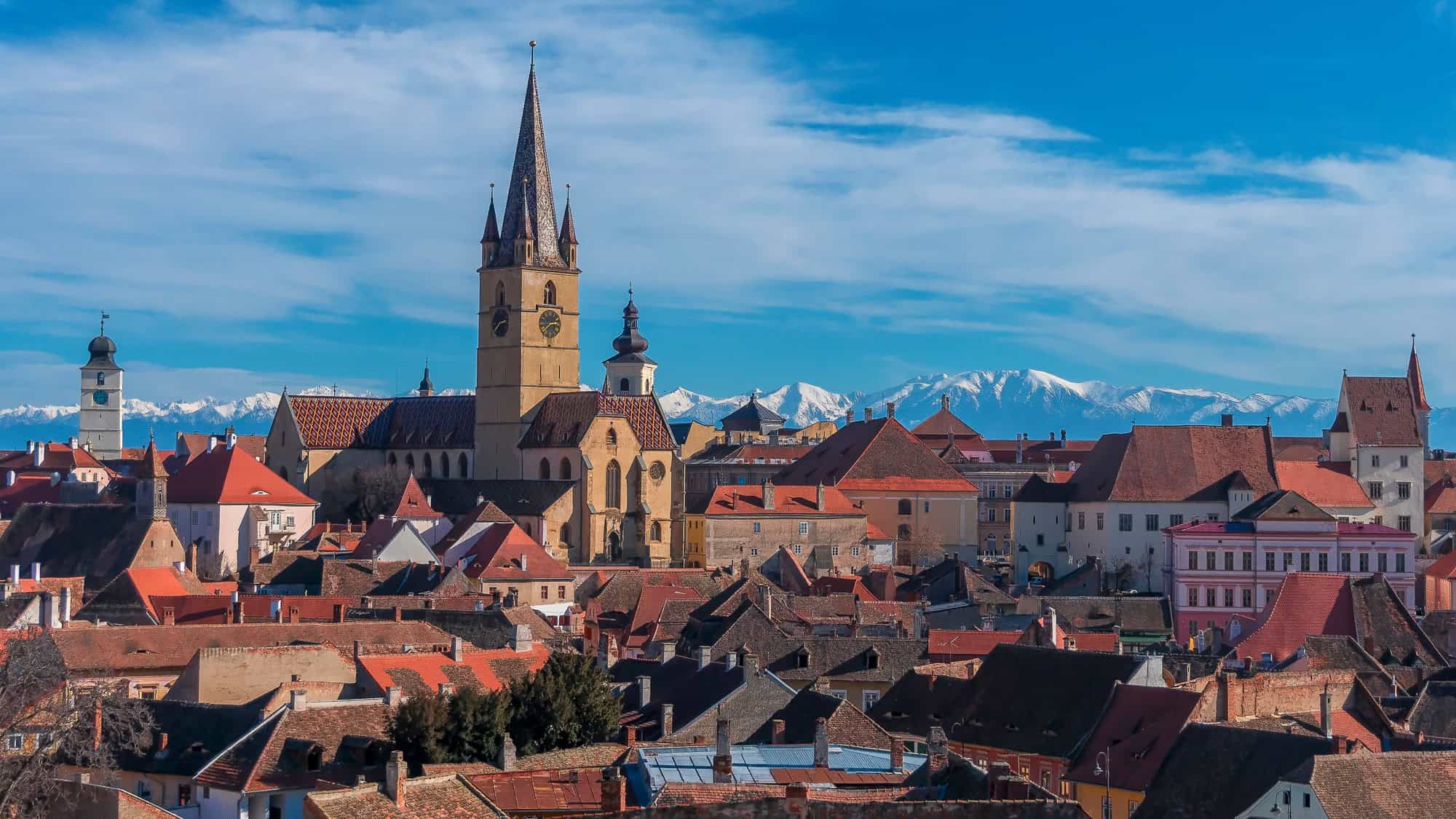 The medieval rooftops and churches of Sibiu, Romania rise against a backdrop of the snow-capped Carpathian Mountains under a crisp blue sky.