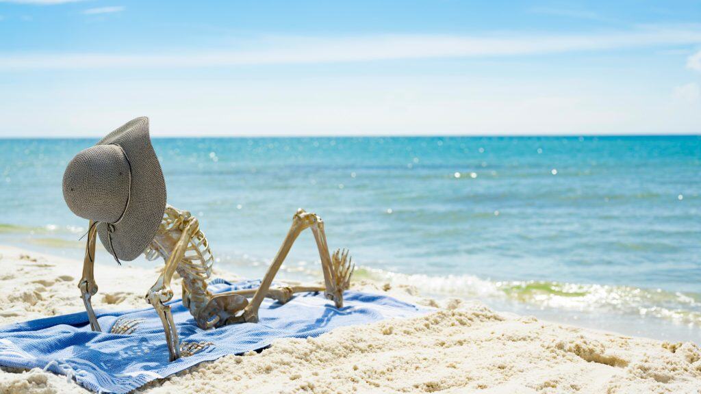 A plastic skeleton wearing a wide-brimmed sunhat lounges on a beach towel facing the ocean, humorously posed as if sunbathing on a sunny shore.