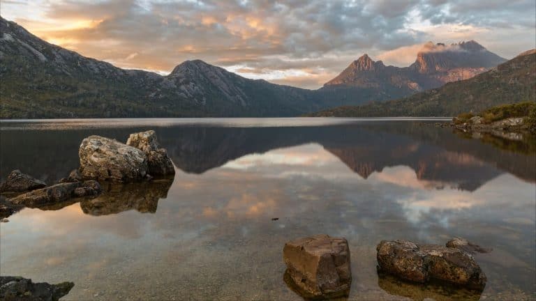 A serene mountain lake reflects the rugged peaks of Cradle Mountain in Tasmania, framed by scattered boulders and a golden sunset sky.