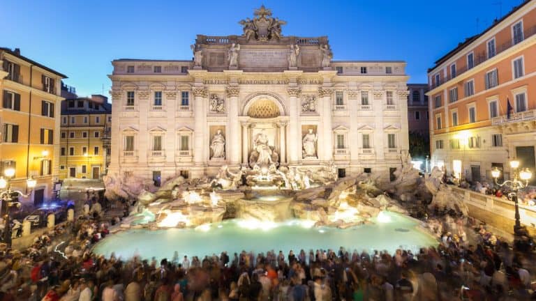 The Trevi Fountain in Rome glows under soft evening lights as a large crowd gathers around its turquoise waters and intricate marble sculptures set against historic buildings.