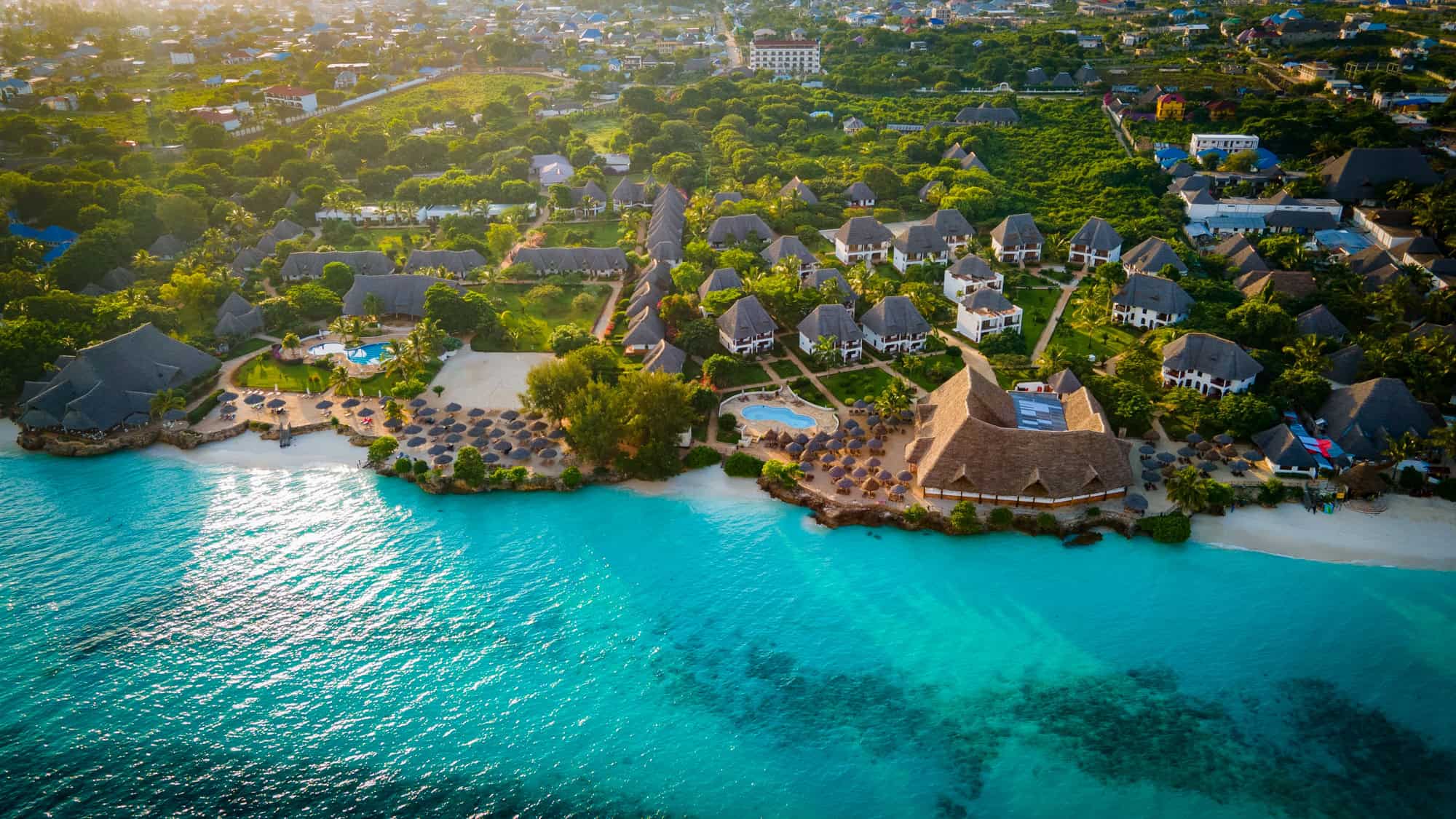 An aerial view of a luxury beachfront resort in Zanzibar shows clusters of thatched-roof villas surrounded by palm trees and vibrant turquoise waters.