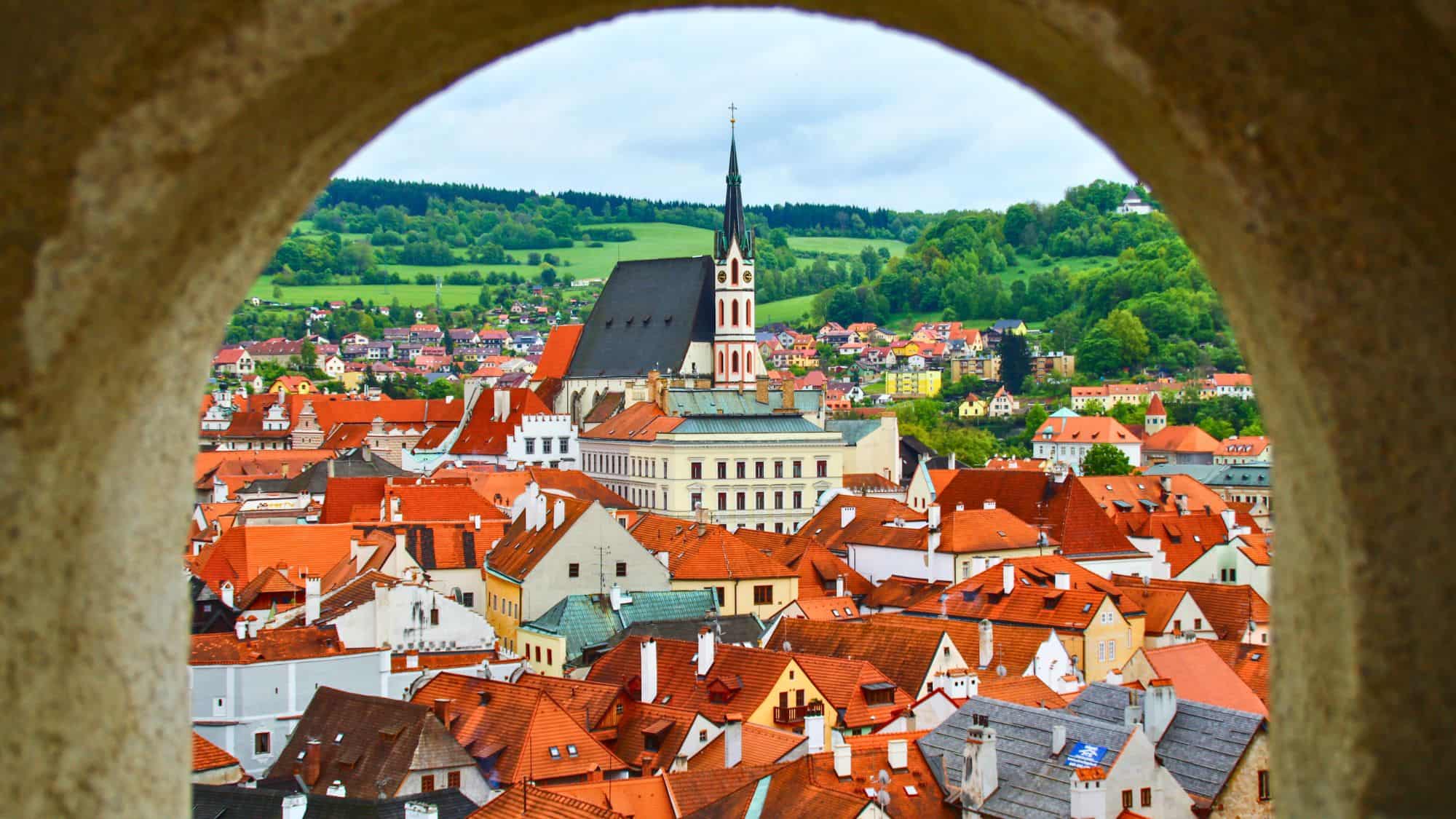 A view through a stone arch reveals a fairytale town of red-roofed buildings clustered around the Gothic-style Church of St. Vitus, with green hills stretching out beyond.