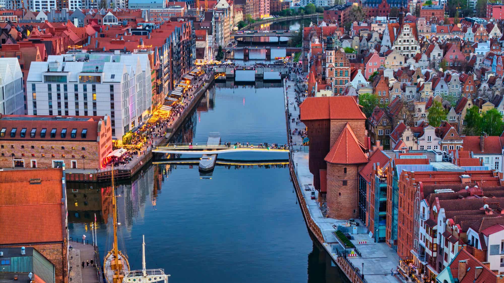 A calm river reflects the blend of modern and historic architecture lining both banks, where cafés and promenades bustle under the soft glow of evening lights.