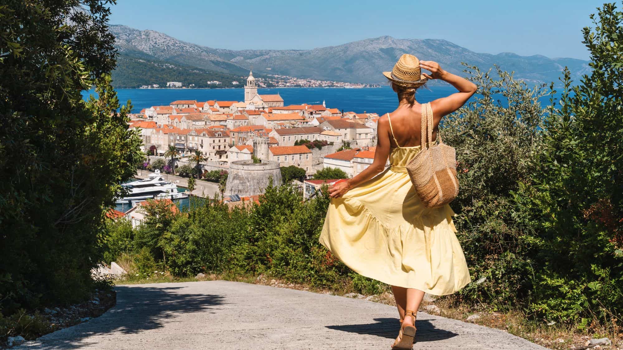 A woman in a flowing yellow dress looks out over the terracotta rooftops and medieval stone walls of an island town nestled between forested hills and deep blue water.