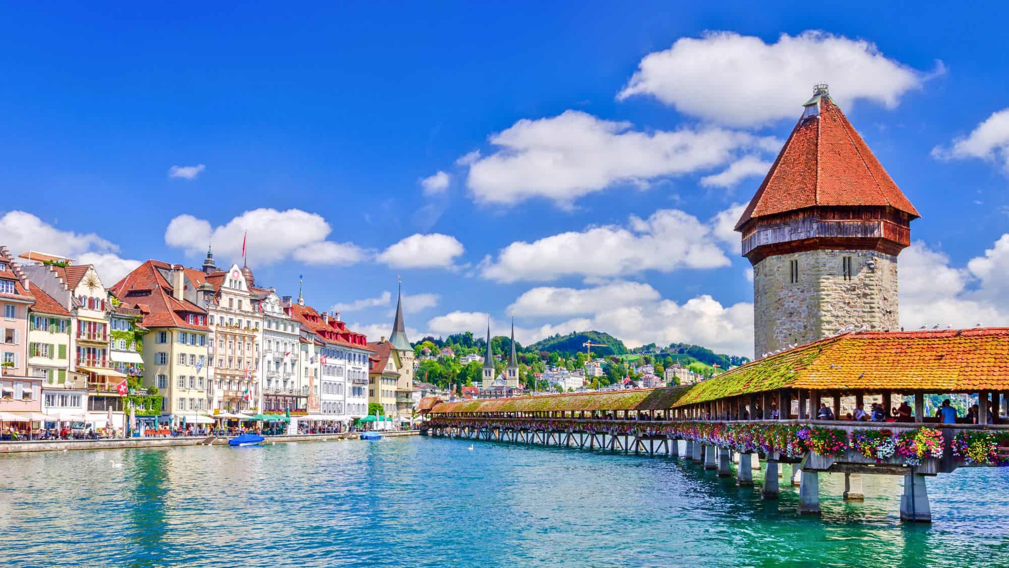 Colorful medieval buildings line the Reuss River beside the iconic Kapellbrücke, a covered wooden bridge decorated with vibrant flower boxes. The scene is framed by clear skies and the stone Water Tower.