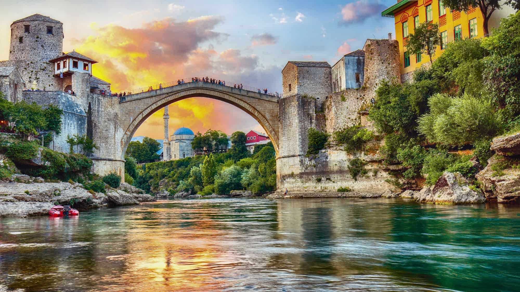 The historic Stari Most arch bridge stretches across the Neretva River, surrounded by stone towers and lush greenery. A glowing sunset casts warm tones across the old town’s skyline.