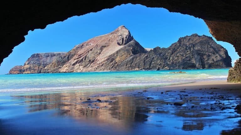 A natural rock arch frames a secluded beach with turquoise waves and a rugged, volcanic peak rising in the distance. The sheltered view highlights the raw, dramatic coastline of Porto Santo Island.