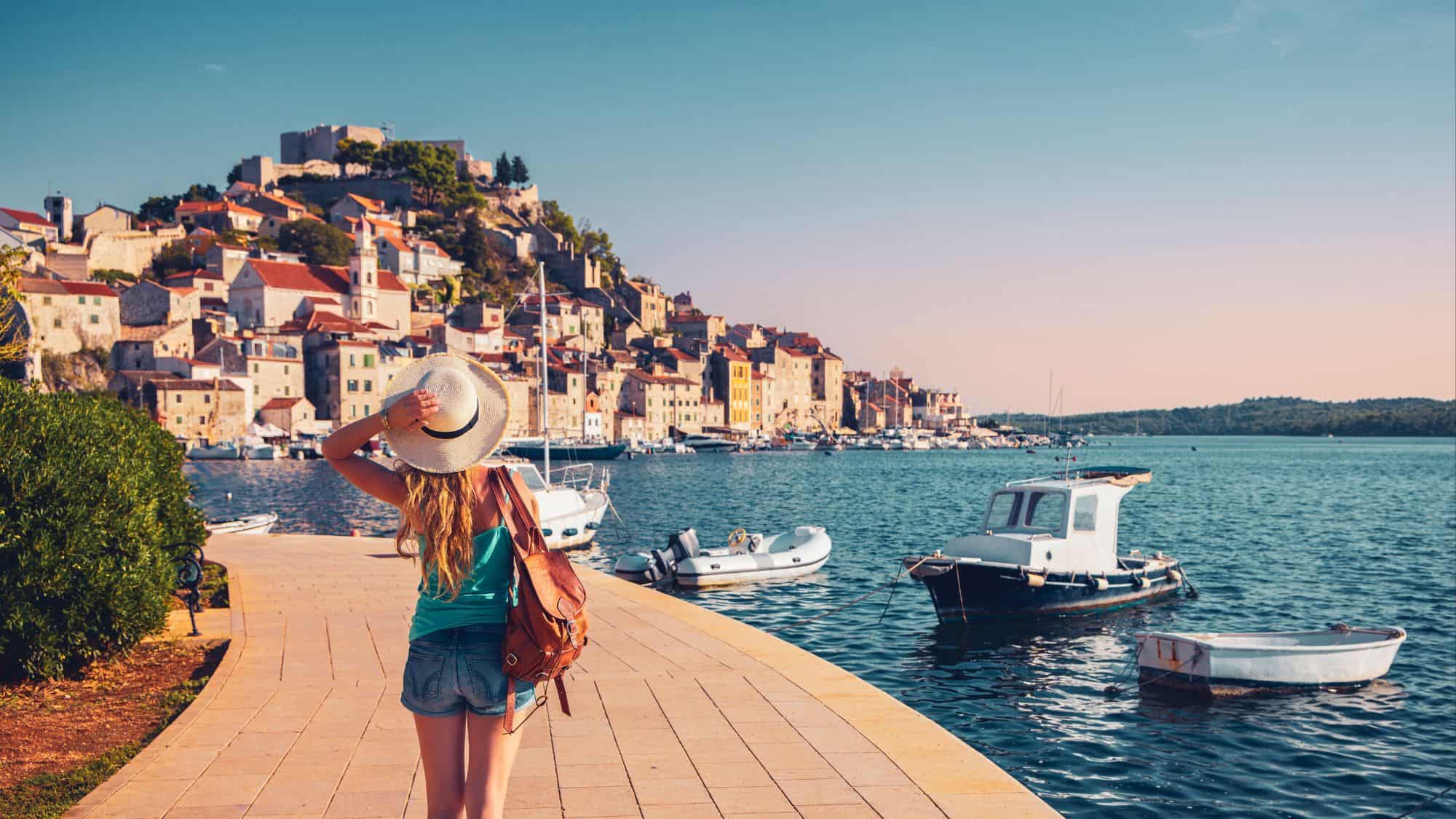 A woman strolls along the promenade looking out toward a coastal town built into the hillside, with boats bobbing gently in the harbor. The sun casts a golden light over the stone buildings and red rooftops.