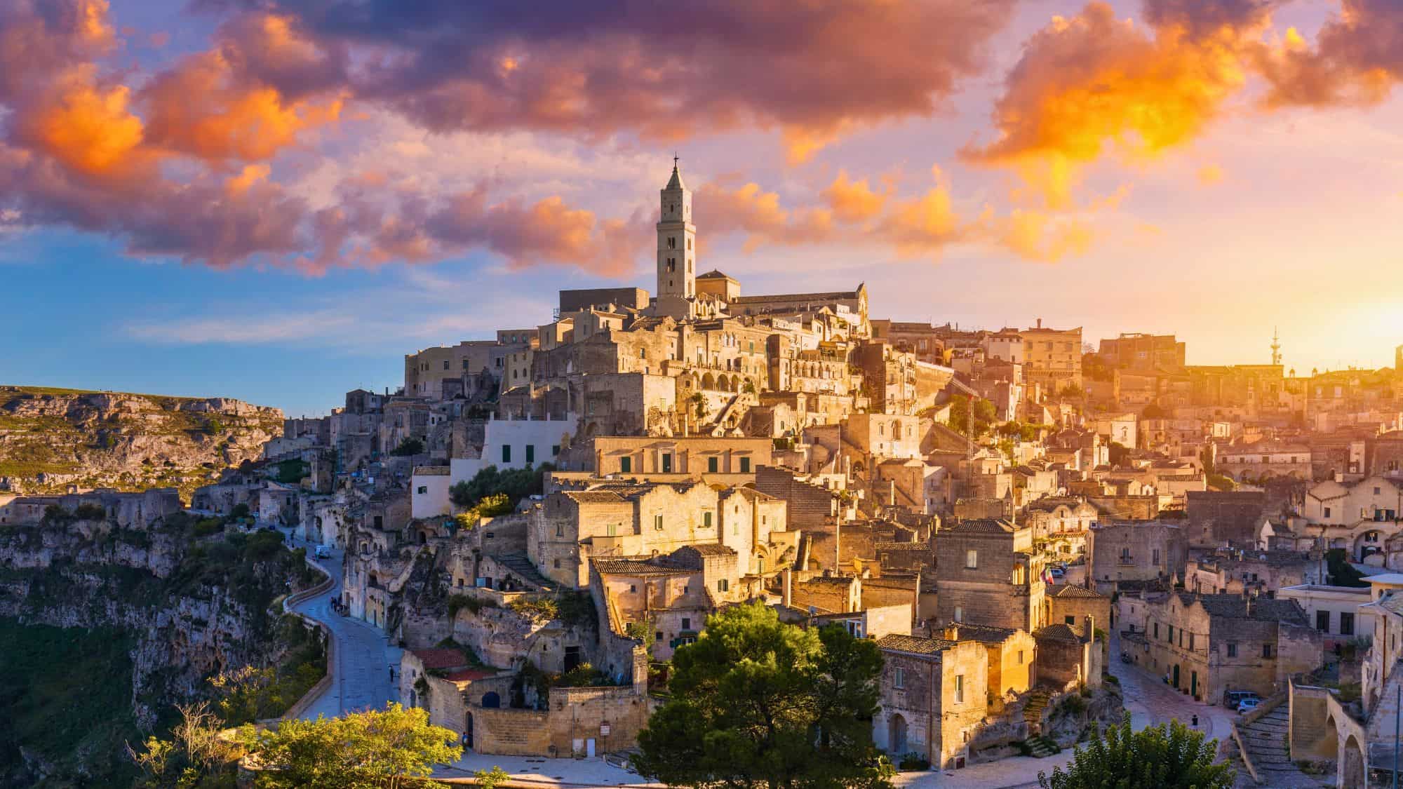 Stone dwellings climb the hillside in layers, culminating in a bell tower that catches the last rays of the day. The town’s ancient cave homes and narrow alleys glow under a fiery sunset sky.