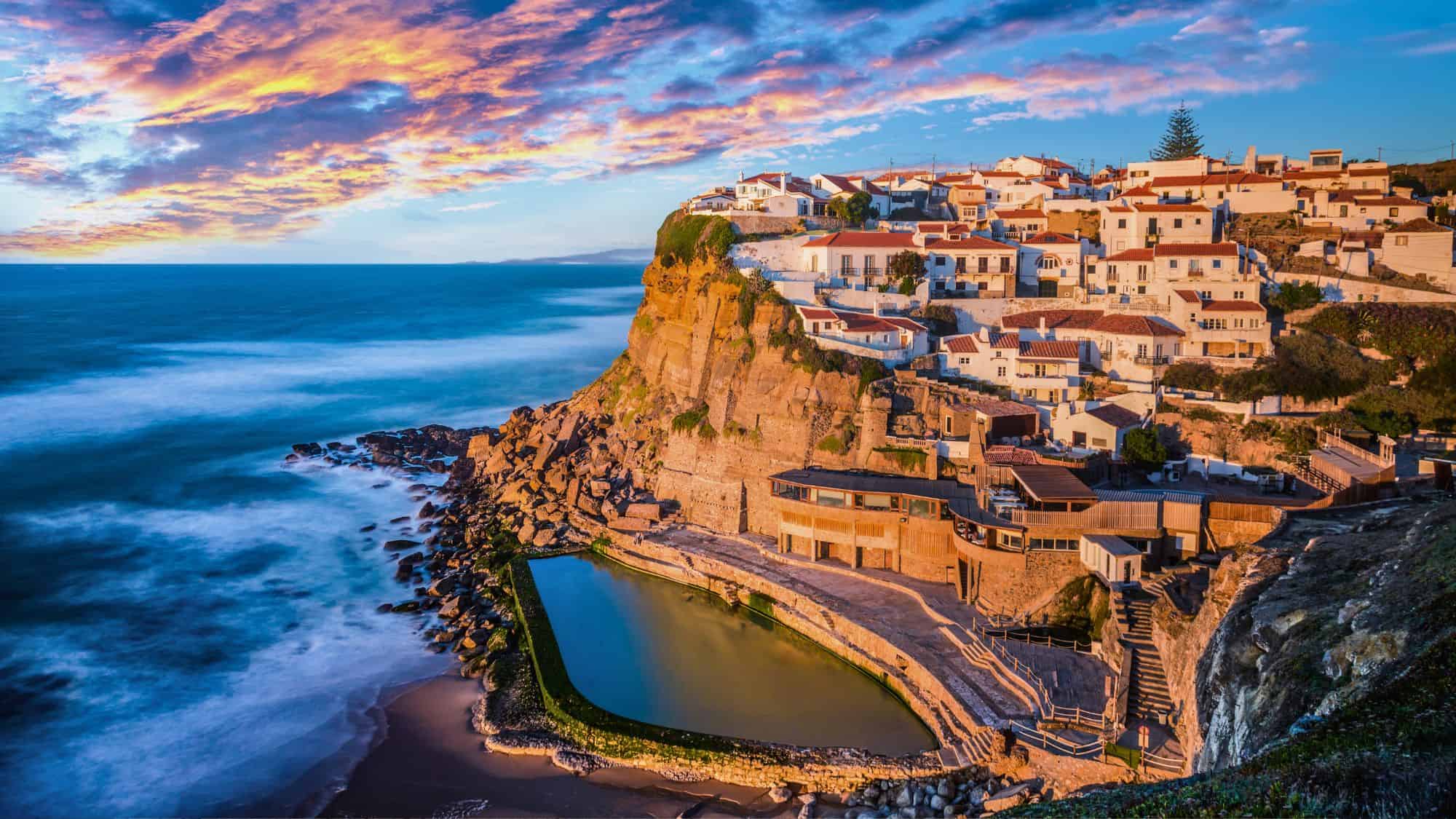 Whitewashed houses perch atop a dramatic cliff, overlooking an ocean pool carved into the rocks below. The evening light sets the scene aglow, with waves crashing against the rugged coastline.