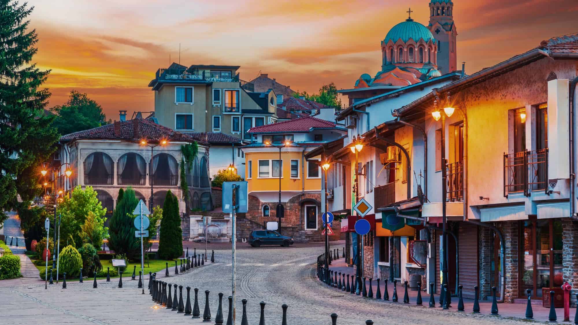 Charming houses with red-tiled roofs and cobbled streets sit beneath the green domes of a church, illuminated by warm streetlights. The town’s layered architecture and historic feel come alive at dusk.