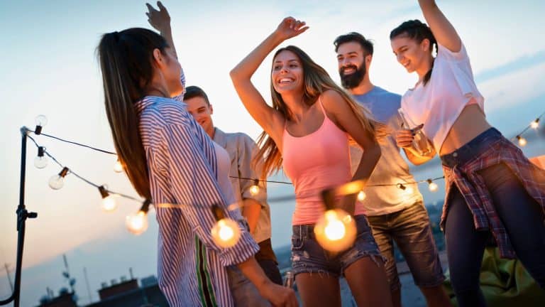 A group of friends dances and laughs under string lights on a rooftop at twilight, lit by the golden glow of sunset and shared energy.