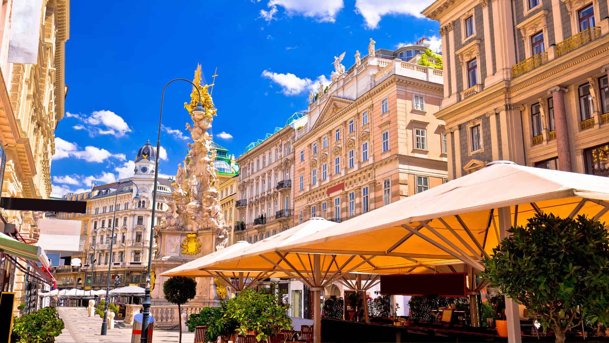 Sunlight pours over grand baroque buildings and a gilded column in a bustling pedestrian square, where café terraces spill out beneath cream-colored umbrellas.