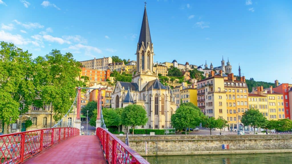 anoramic view of Lyon's riverside with the red suspension bridge, Saint-Georges Church, and colorful hillside buildings under a clear sky.