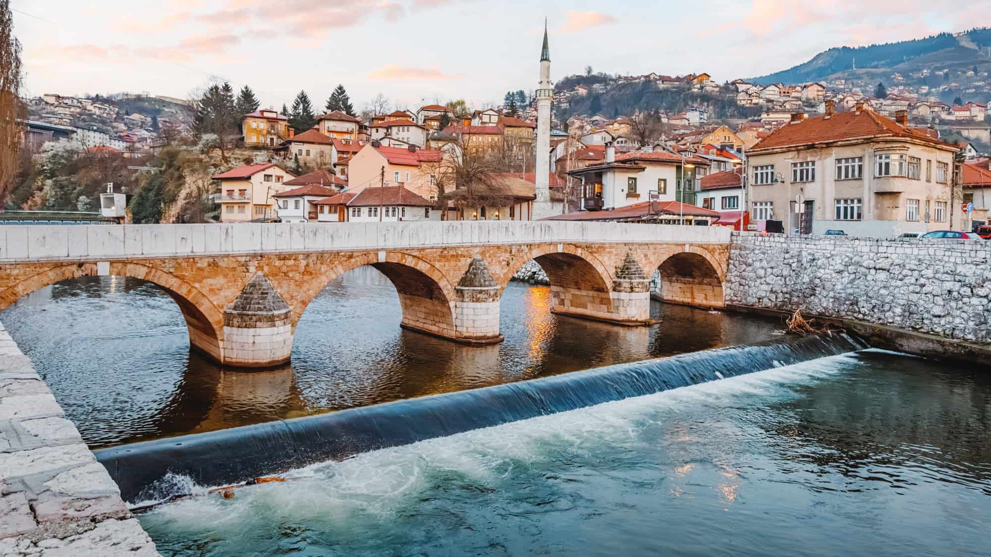 Scenic view of Sarajevo at sunset, featuring a historic stone bridge, cascading river, and a hillside mosque with a prominent minaret.